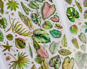 May include: A sheet of gold foil stickers featuring various tropical leaves and plants. The stickers are on a clear background and have a glossy finish.