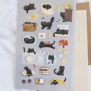Könnte beinhalten: Bogen mit dekorativen Aufklebern mit Illustrationen von schwarzen Katzen in verschiedenen Posen. Die Aufkleber zeigen Katzen mit Garn, in einer Kaffeetasse und die hinter einer Pflanze hervorlugen. Der Bogen trägt die Aufschrift "DAILY DECO STICKERS" und "my little universe".