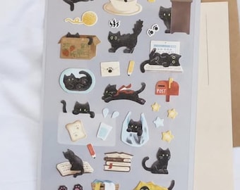 Puede incluir: Hoja de pegatinas decorativas con ilustraciones de gatos negros en varias poses. Las pegatinas incluyen gatos con hilo, en una taza de café y asomándose detrás de una planta. La hoja está etiquetada como "DAILY DECO STICKERS" y "my little universe".