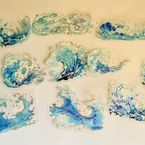 May include: A selection of wave-shaped stickers in different sizes. The stickers are in shades of blue and white with iridescent highlights, giving a shimmering appearance. The designs represent ocean waves in a stylized, artistic style.