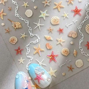May include: A sheet of nail art stickers featuring various seashells and starfish in shades of beige, red, and iridescent colors. Two painted nails with a blue, pink, and white design are also visible, with a starfish and seashell theme.