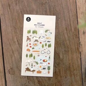 May include: A sheet of colorful stickers featuring illustrations of a picnic in a garden. The stickers include images of trees, a table, chairs, food, drinks, a bicycle, a beach umbrella, a lounge chair, and other items. The sheet is labeled "Daily Deco Stickers" and "Daily - Picnic Garden".