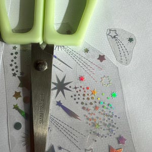 May include: A pair of green-handled scissors rests on a sheet of clear stickers with colorful star designs. The scissors are stainless steel. The stickers feature various sizes and colors of stars, including holographic and rainbow effects.