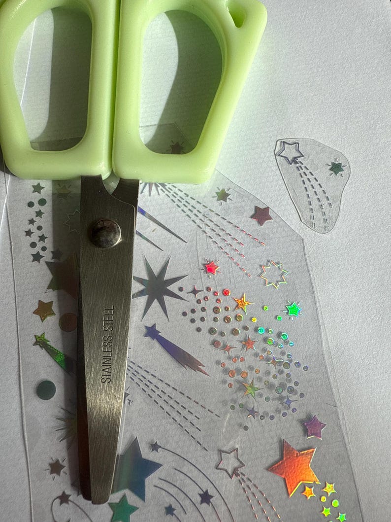 May include: A pair of green-handled scissors rests on a sheet of clear stickers with colorful star designs. The scissors are stainless steel. The stickers feature various sizes and colors of stars, including holographic and rainbow effects.