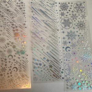 May include: Three clear sticker sheets with holographic designs. The designs include stars, snowflakes, shooting stars, and crescent moons. The stickers have a metallic sheen and are arranged in vertical strips.