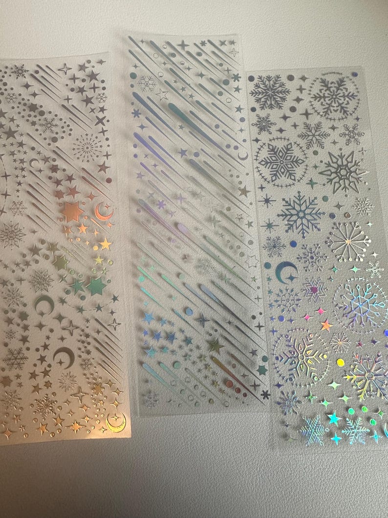 May include: Three clear sticker sheets with holographic designs. The designs include stars, snowflakes, shooting stars, and crescent moons. The stickers have a metallic sheen and are arranged in vertical strips.