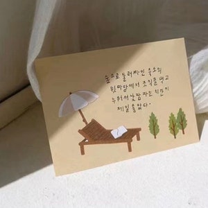 May include: A beige card with a handwritten message in Korean. The card features a cartoon illustration of a beach scene with a lounge chair, an umbrella, and two trees. The text on the card reads: "숲으로 둘러싸인 숙소의 뒷마당에서 조식을 먹고 누워서 낮잠자는 시간이 제일좋았다."