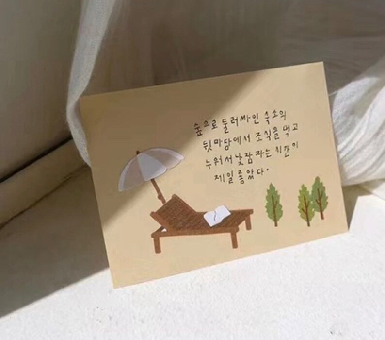 May include: A beige card with a handwritten message in Korean. The card features a cartoon illustration of a beach scene with a lounge chair, an umbrella, and two trees. The text on the card reads: "숲으로 둘러싸인 숙소의 뒷마당에서 조식을 먹고 누워서 낮잠자는 시간이 제일좋았다."