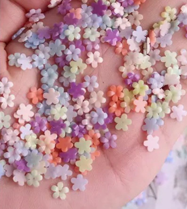 K&ouml;nnte beinhalten: Eine Sammlung von pastellfarbenen, blumenf&ouml;rmigen Streuseln. Die Streusel sind in verschiedenen Farben, darunter rosa, lila, blau und gelb.