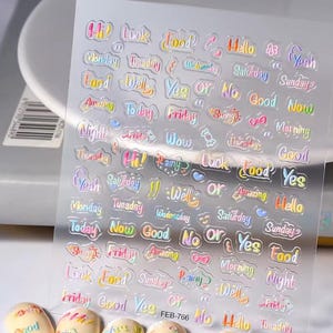 May include: Clear sheet of nail art stickers with colorful text including words like "Hello", "Food", and days of the week. The stickers have a holographic, iridescent finish. The sheet is labeled "FEB-766".