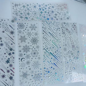 May include: Clear sticker sheets with celestial designs. The sheets feature silver and iridescent stars, snowflakes, shooting stars, and crescent moons. The stickers are arranged in vertical strips and a single rectangular sheet.