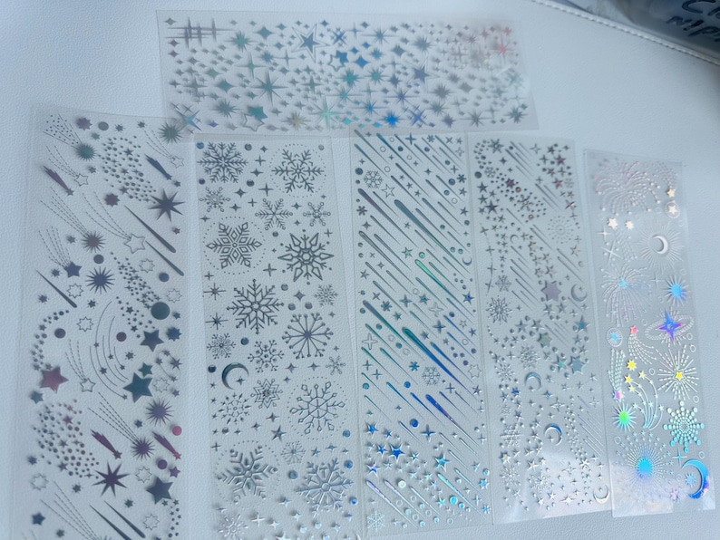 May include: Clear sticker sheets with celestial designs. The sheets feature silver and iridescent stars, snowflakes, shooting stars, and crescent moons. The stickers are arranged in vertical strips and a single rectangular sheet.