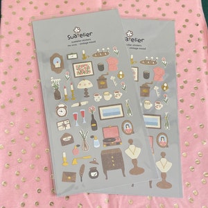 May include: Two sheets of stickers with a vintage aesthetic. The stickers feature illustrations of various objects, including a coffee pot, a clock, a key, a bottle of wine, a record player, a lamp, a vase, a necklace, and a quill pen. The stickers are on a grey background with the text "Sua*telier" and "no.3133 vintage mood" on one sheet and "atelier" and "vintage mood" on the other sheet.