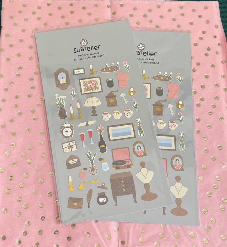 May include: Two sheets of stickers with a vintage aesthetic. The stickers feature illustrations of various objects, including a coffee pot, a clock, a key, a bottle of wine, a record player, a lamp, a vase, a necklace, and a quill pen. The stickers are on a grey background with the text "Sua*telier" and "no.3133 vintage mood" on one sheet and "atelier" and "vintage mood" on the other sheet.