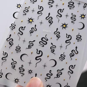 May include: A sheet of nail art stickers featuring black snakes, gold crescent moons, gold suns with central gems, and small gold star accents. The sheet is labelled with the text 5D-0476 and contains multiple individual adhesive designs.