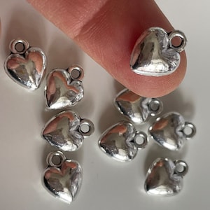 Op de afbeelding: Een groep van acht zilveren hartvormige bedels met een lus aan de bovenkant. De bedels zijn verspreid over een wit oppervlak.