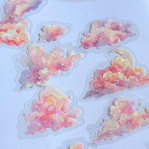 Puede incluir: Una colección de pegatinas con forma de nube en tonos rosa, naranja y morado. Las pegatinas tienen un efecto brillante e iridiscente, algunas con estrellas y una luna creciente. Sobre un fondo blanco.