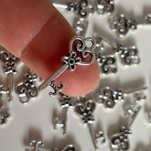 Op de afbeelding: Een close-up van een zilveren sleutelbedel met een decoratief hartvormig hoofd. De sleutel is omringd door veel andere vergelijkbare bedels.