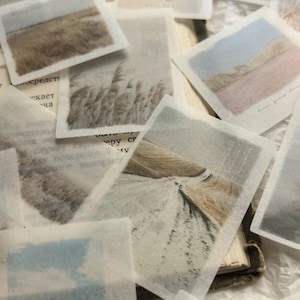 Puede incluir: Una colección de pequeñas pegatinas cuadradas y translúcidas con escenas de paisajes. Las imágenes representan campos, agua y cielos en tonos apagados de marrón, azul y beige. Algunas pegatinas están sobre páginas de libros abiertos.