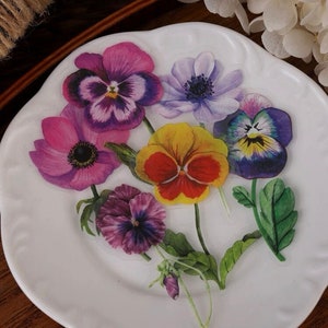 May include: A white plate with a floral design featuring five flowers. The flowers are a mix of pink, purple, yellow, and blue. The flowers are arranged in a bouquet with green leaves.