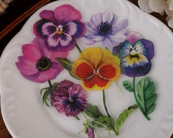 May include: A collection of colorful floral stickers featuring pansies and anemones. The stickers are arranged on a white, scalloped-edge plate. The flowers display shades of purple, pink, yellow, and red, with green leaves.
