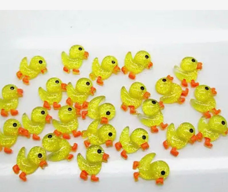K&ouml;nnte beinhalten: Eine Sammlung von gelben Glitzer-Enten-Anh&auml;ngern mit orangefarbenen Schn&auml;beln und F&uuml;&szlig;en. Die Anh&auml;nger sind perfekt, um Kunsthandwerk, Schmuck und anderen Projekten einen Hauch von Verspieltheit zu verleihen.