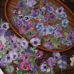 May include: A set of floral stickers featuring purple, blue, and pink flowers with green leaves. The stickers are on a wooden surface.