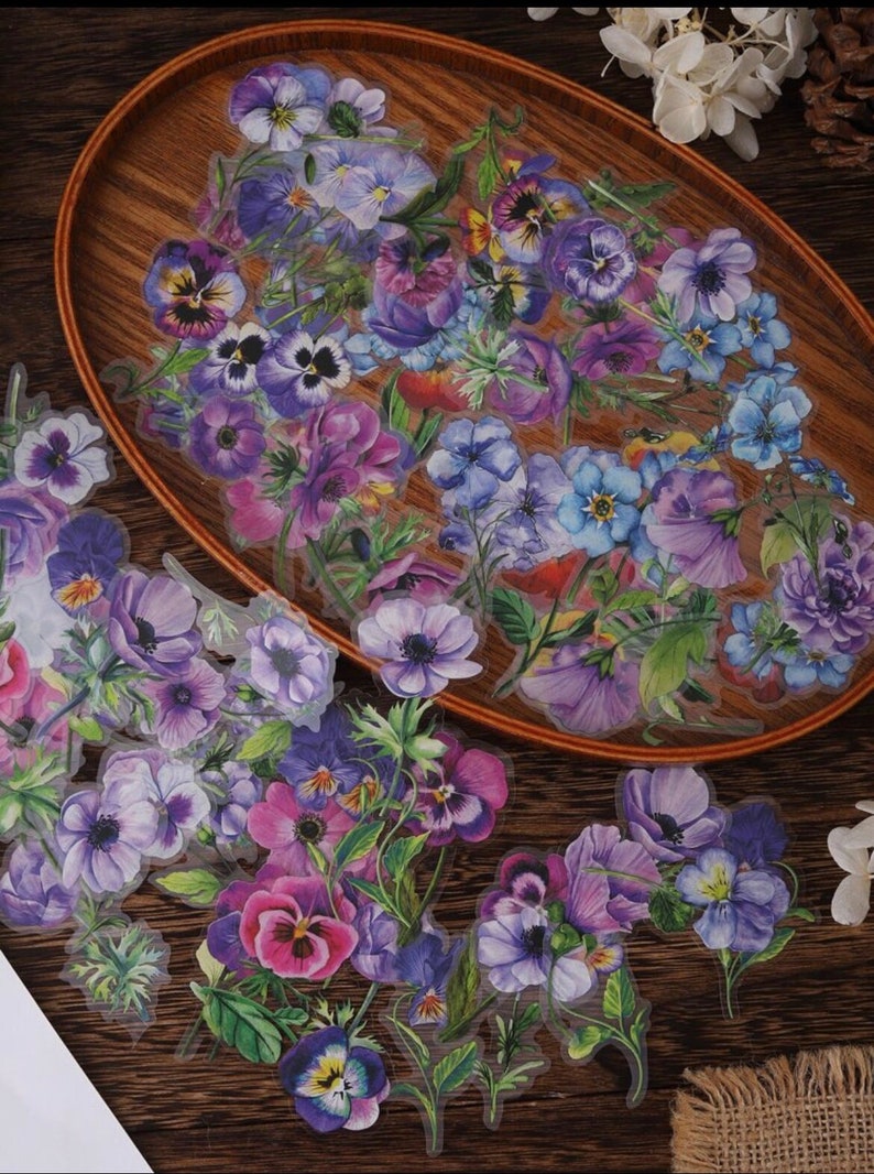 May include: A set of floral stickers featuring purple, blue, and pink flowers with green leaves. The stickers are on a wooden surface.