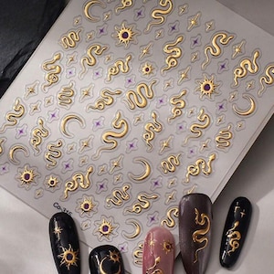 May include: A sheet of gold nail art stickers with snake, moon, star, and sun designs. Several painted fingernails are displayed below, showcasing the nail art designs. The nails are painted in various colours, including black, brown, and pink.