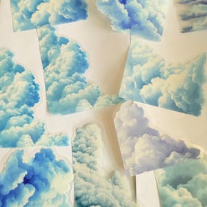 May include: A collection of cloud-shaped stickers in various sizes. The stickers feature a realistic depiction of fluffy white and blue clouds against a light background. The stickers are cut out and ready to use.