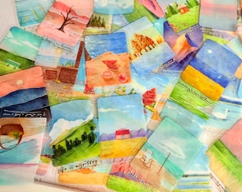 May include: A collection of watercolor art cards featuring various landscape scenes. The cards depict beaches, fields, trees, and buildings, with soft colors and handwritten text accents. The cards are arranged in a scattered pattern.
