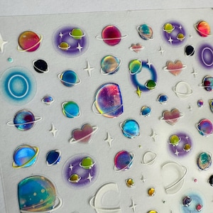 Puede incluir: Hoja de pegatinas holográficas con planetas coloridos, estrellas y planetas en forma de corazón. Las pegatinas tienen un acabado brillante y son en tonos de rosa, azul, verde y morado. Los planetas tienen anillos y estrellas.