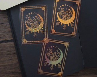 May include: Three gold foil stickers with a crescent moon design. Each sticker features a crescent moon with a cluster of crystals and flowers inside. The stickers have a black background and a gold border.