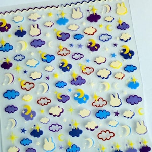 May include: A sheet of stickers featuring a whimsical night sky theme. The stickers include crescent moons, stars, clouds, and bunny silhouettes in shades of blue, purple, yellow, and white. The stickers are arranged on a clear, wavy-edged sheet.