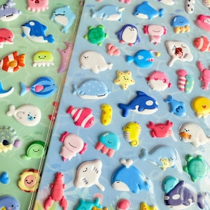 Könnte beinhalten: Zwei Bögen mit bunten, aufgeplusterten Aufklebern mit verschiedenen Meeresbewohnern. Die Aufkleber zeigen Fische, Wale, Delfine, Seesterne und andere Meereslebewesen in Blau-, Rosa-, Gelb- und Grüntönen. Die Aufkleber sind auf einem hellblauen und grünen Hintergrund angeordnet.