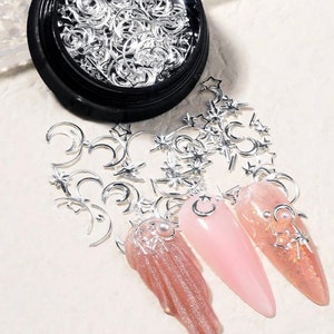 Könnte beinhalten: Ein Behälter mit silbernen Nagelkunst-Anhängern in Form von Monden und Sternen. Die Anhänger sind auf einer weißen Oberfläche verstreut, auf der drei rosafarbene Nagelspitzen mit den Anhängern verziert sind.