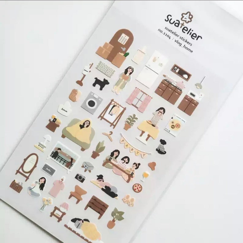 K&ouml;nnte beinhalten: Eine Bogen mit Aufklebern, die Illustrationen eines Hausinneren zeigen, darunter M&ouml;bel, Ger&auml;te und Personen. Die Aufkleber sind in verschiedenen Farben gehalten, darunter Braun, Beige und Rosa. Der Bogen ist mit "Suatelier Stickers no.1104: Vlog Home" beschriftet.