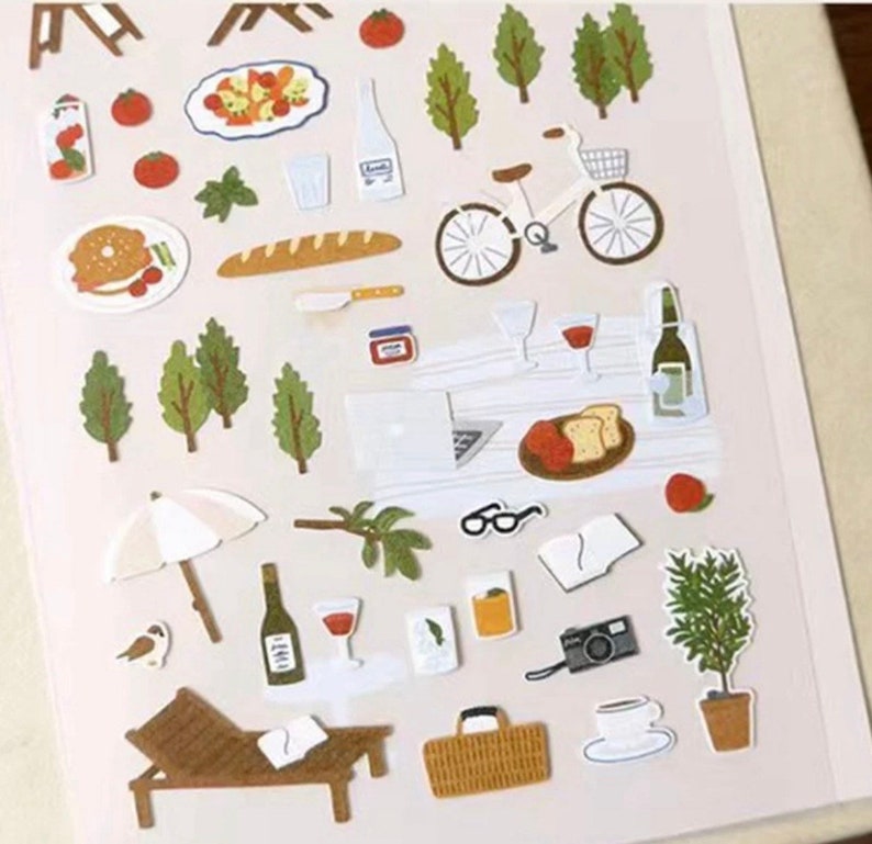 May include: A sheet of colorful stickers featuring various picnic and outdoor themed illustrations. The stickers include a bicycle, trees, a baguette, a picnic basket, a glass of wine, a camera, a lounge chair, and more.
