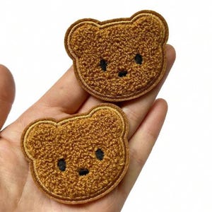 Pode incluir: Dois emblemas em forma de urso castanho com uma superfície texturizada e felpuda. Cada emblema apresenta detalhes bordados, incluindo dois pequenos olhos circulares pretos e um nariz preto minúsculo, contornados por uma costura castanha.