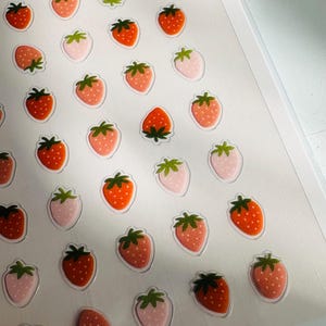May include: A sheet of stickers featuring a repeating pattern of pink and orange strawberries. The stickers are on a white background and have a glossy finish. The sheet is labeled "DECO" in the bottom right corner.
