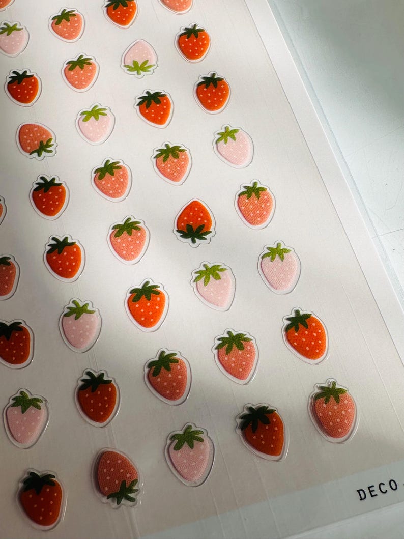May include: A sheet of stickers featuring a repeating pattern of pink and orange strawberries. The stickers are on a white background and have a glossy finish. The sheet is labeled "DECO" in the bottom right corner.