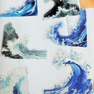 Puede incluir: Una colección de pegatinas con forma de ola en tonos azules, blancos y grises. Las pegatinas representan olas oceánicas con detalles intrincados, capturando el movimiento y la textura del agua. Algunas pegatinas tienen un borde transparente.