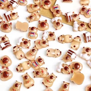 May include: A selection of small, tan bear-shaped resin charms with brown detailing. Some charms feature additional elements such as a small cake or a drink. The charms are spread across a white surface.