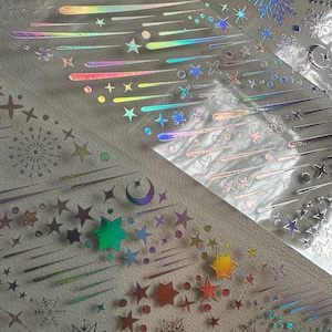 May include: Iridescent stickers featuring celestial designs. The stickers include stars, shooting stars, moons, and snowflakes in a range of colors. The stickers are arranged on a light-colored surface, creating a shimmering effect.