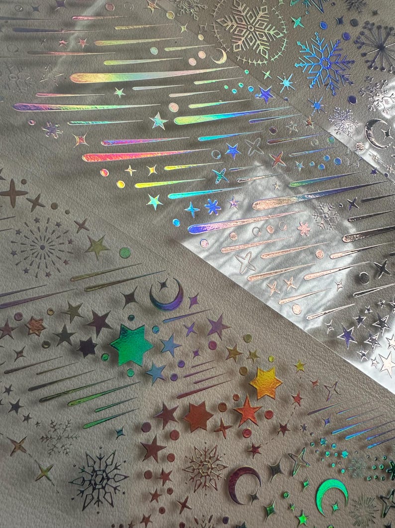 May include: Iridescent stickers featuring celestial designs. The stickers include stars, shooting stars, moons, and snowflakes in a range of colors. The stickers are arranged on a light-colored surface, creating a shimmering effect.