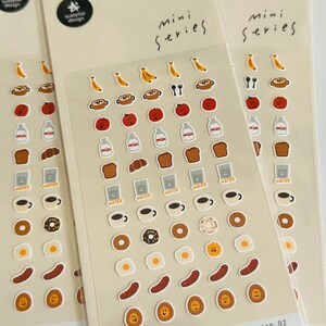 Può includere: Tre fogli di adesivi con illustrazioni a tema cibo. Ogni foglio presenta adesivi di banane, mele, bottiglie di latte, pane, tazze di caffè, ciambelle e uova fritte. Il testo "mini series" e "FOOD.02" sono visibili.