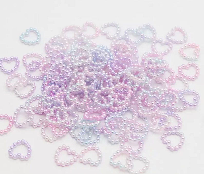 K&ouml;nnte beinhalten: Ein gro&szlig;er Haufen aus schimmernden, pastellfarbenen, rosa, lila und blauen Perlen in Herzform.