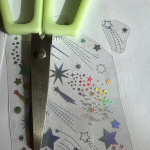 May include: A pair of light green handled scissors cutting a sheet of holographic star stickers. The stickers feature various star designs and shooting star trails. The scissors are stainless steel.