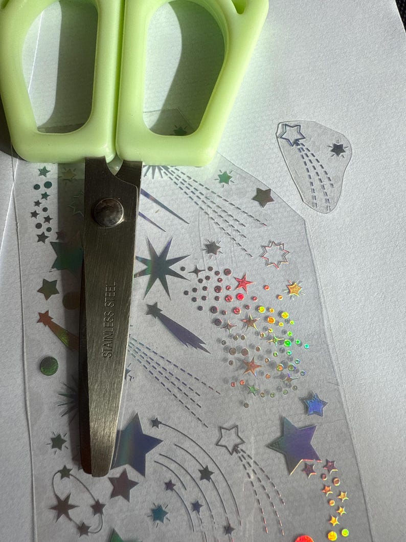 May include: A pair of light green handled scissors cutting a sheet of holographic star stickers. The stickers feature various star designs and shooting star trails. The scissors are stainless steel.