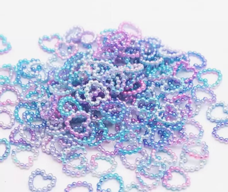 K&ouml;nnte beinhalten: Ein gro&szlig;er Haufen regenbogenfarbener herzf&ouml;rmiger Perlen in Blaut&ouml;nen, Rosa und Lila. Die Perlen sind aus Kunststoff und haben eine glatte, gl&auml;nzende Oberfl&auml;che.